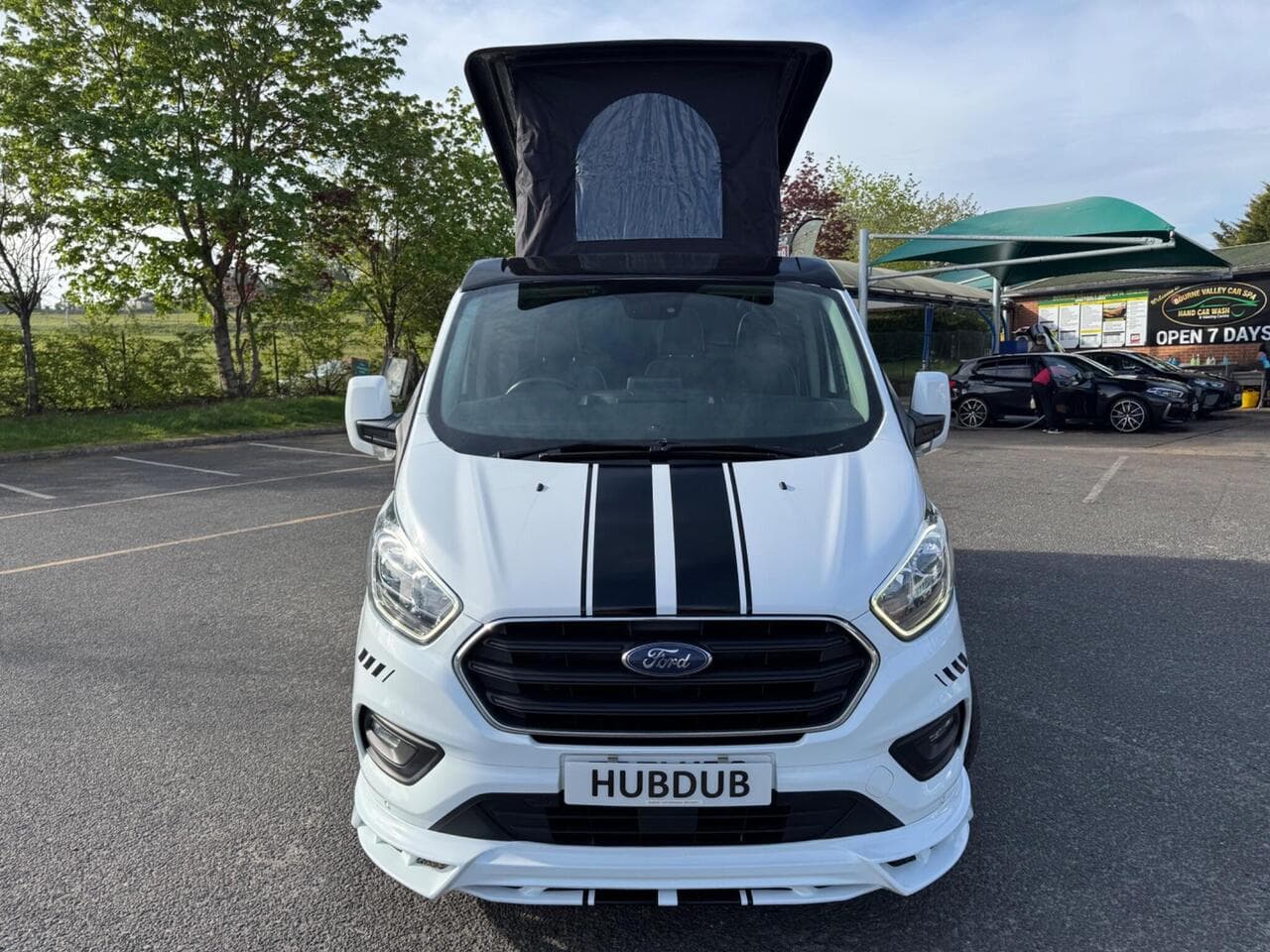 A HubDub-converted Ford Transit Custom in white with twin black bonnet stripes and its pop top raised, photographed head-on