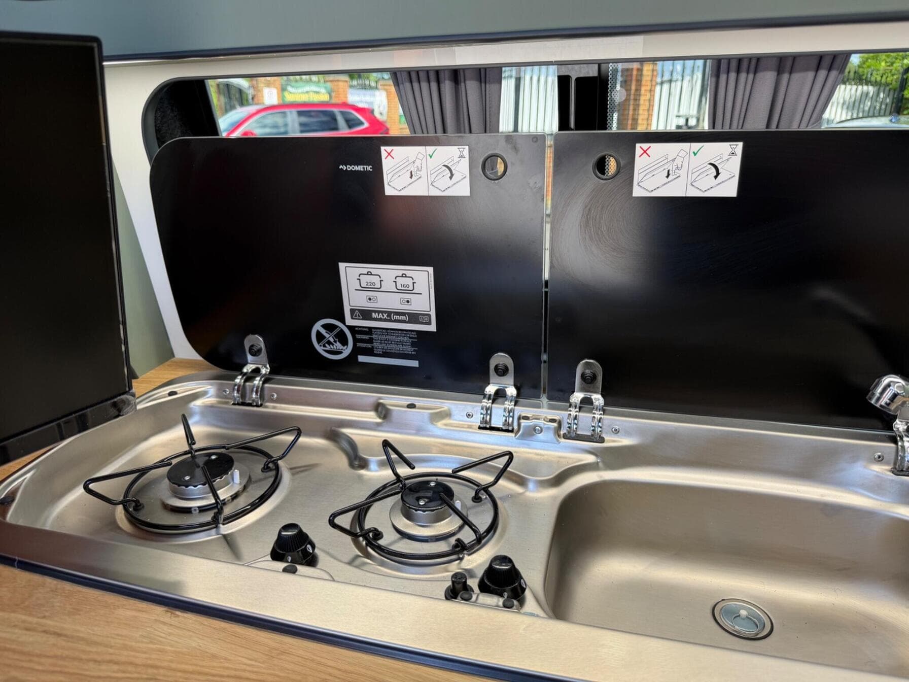 Dometic two-burner gas hob and stainless steel sink combination unit set into an oak worktop, with the glass lids raised