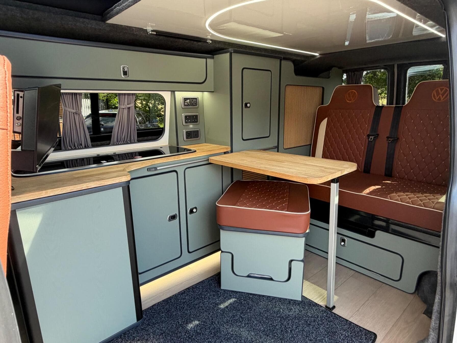 Interior of a HubDub-converted 2021 VW Transporter T6.1 campervan, showing the sage green galley, oak worktop, fold-away table and rust leather rock and roll bed