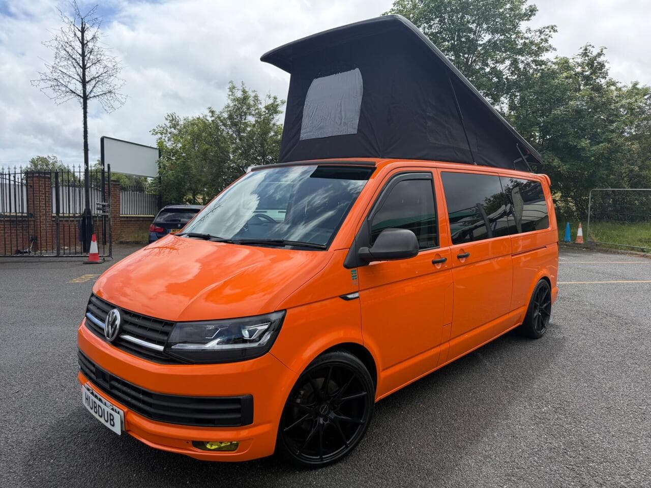 A HubDub-converted VW Transporter T6 in orange with its gloss black pop top elevated, photographed three-quarter front